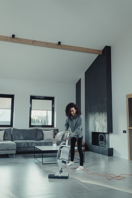 A professional in full protective gear, including a white coverall, face mask, and blue gloves, is performing deep cleaning and sanitisation on a beige carpet in a modern living room. The worker is us