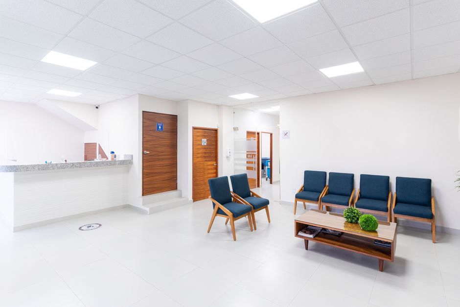 A clean, well-lit hospital corridor with white tiled flooring and cream walls featuring some framed notices. On the left side are several white and gray striped chairs neatly arranged along the wall,