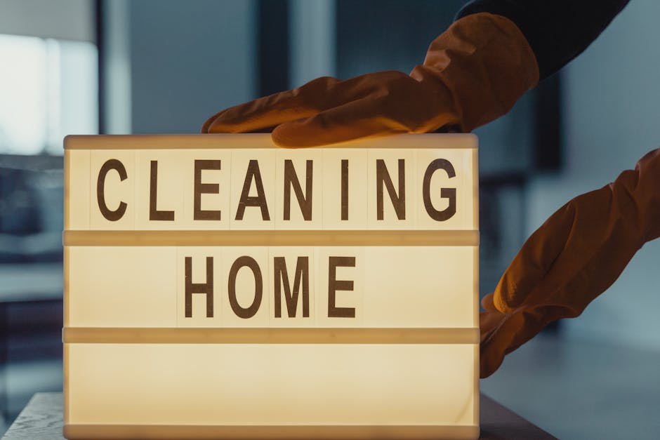 A beige or taupe painted wall in a residential or commercial space with a small blue plastic or silicone wall hook hanging a wooden-handled cleaning brush with natural bristles. The hook is mounted to